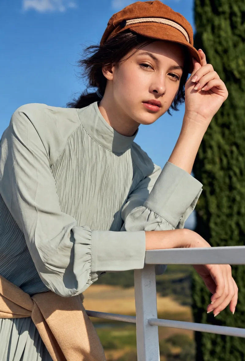 Natural-light portrait of a model in an orange cap and sage blouse beside a railing