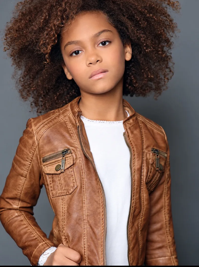 Young performer portrait with curly hair and a brown leather jacket against a gray background